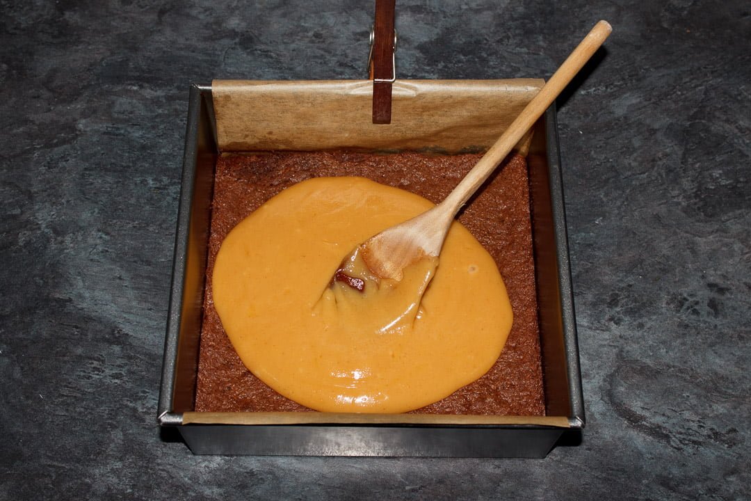 Caramel being spread over brownies with a wooden spoon in a lined square baking tin