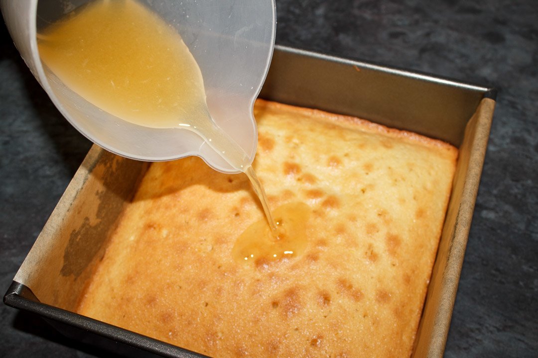 A lemon and sugar mixture being drizzled over the top of a lemon traybake cake