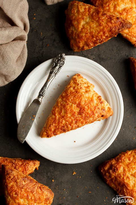 A cheese scone on a white plate with a knife surrounded by a cloth and more cheese scones