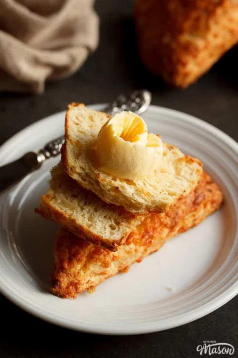 Broken cheese scones on top of a whole cheese scone topped with butter, on a white plate with a knife