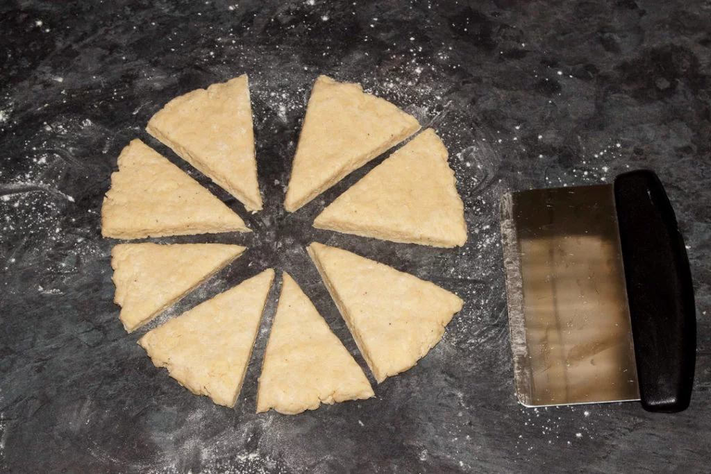 Cheese scones dough cut into triangles with a metal dough scraper on a lightly floured work surface.