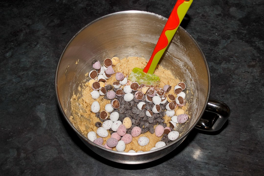 Cookie dough with mini eggs and chocolate chips in a large bowl with a green and orange spatula
