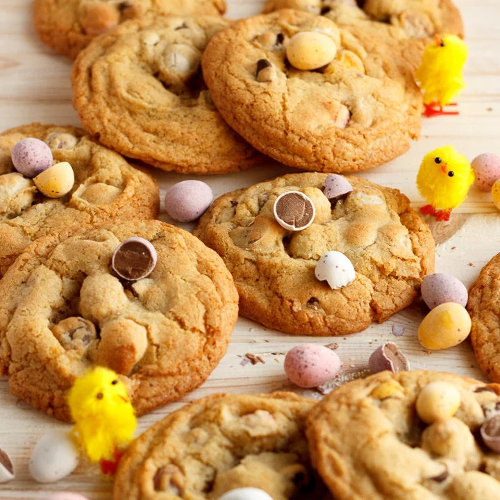 Mini Egg cookies in a pile on a worktop alongside scattered Mini Eggs and little yellow chicks