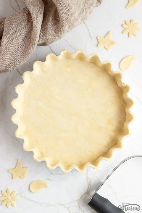 A loose bottomed fluted tart tin lined with unbaked shortcrust pastry surrounded by pastry leaves, a pastry blender and some fabric.