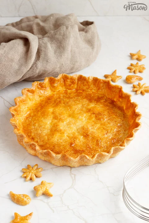 A golden baked shortcrust pastry case surrounded by golden pastry leaves, a pastry blender and a piece of fabric.
