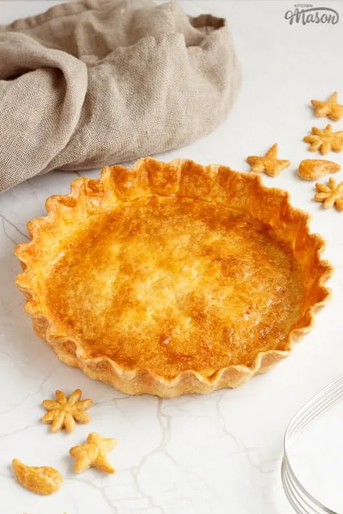 A golden baked shortcrust pastry case surrounded by golden pastry leaves, a pastry blender and a piece of fabric.