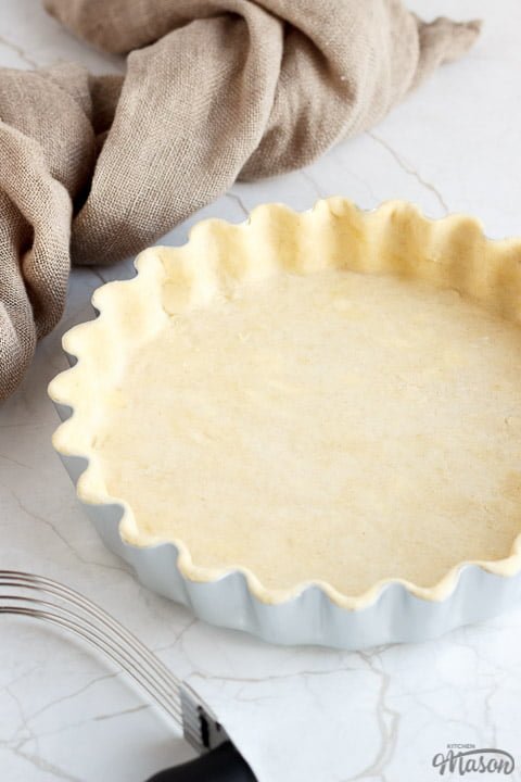 A loose bottomed fluted tart tin lined with unbaked shortcrust pastry surrounded by pastry leaves, a pastry blender and some fabric.