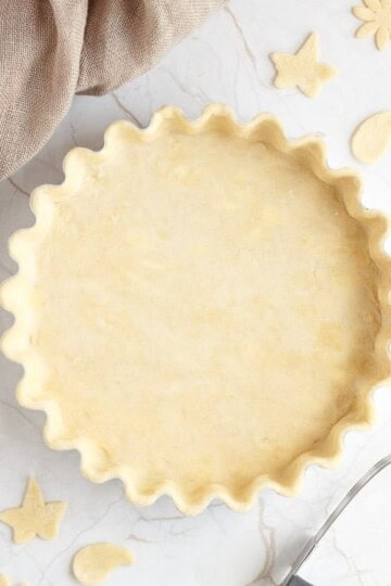 A loose bottomed fluted tart tin lined with unbaked shortcrust pastry surrounded by pastry leaves, a pastry blender and some fabric.
