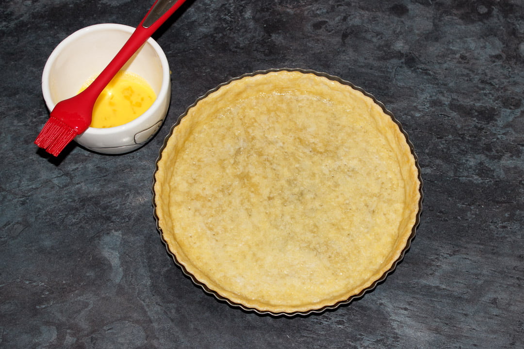 Part blind baked shortcrust pastry case in a round fluted tart tin that's been brushed with beaten egg on a work surface