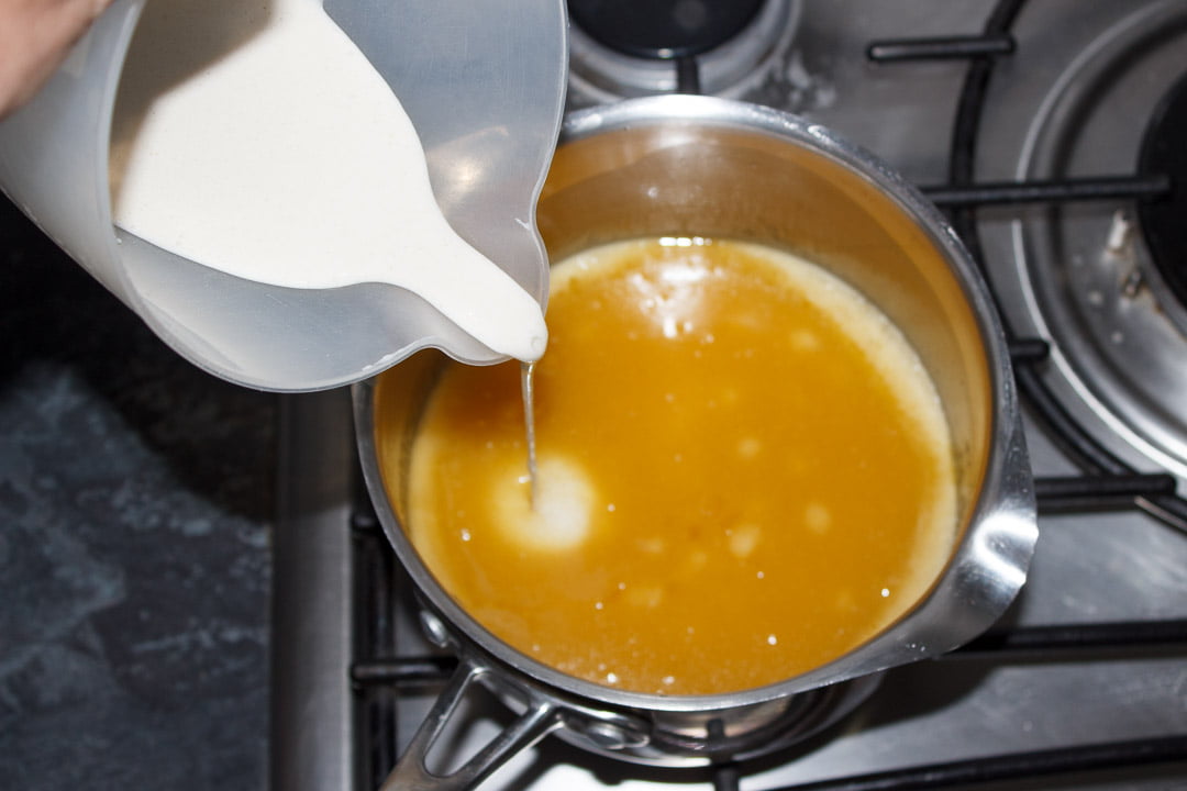 Milk and flour paste being poured into a melted butterscotch tart filling in a large saucepan on a stove top