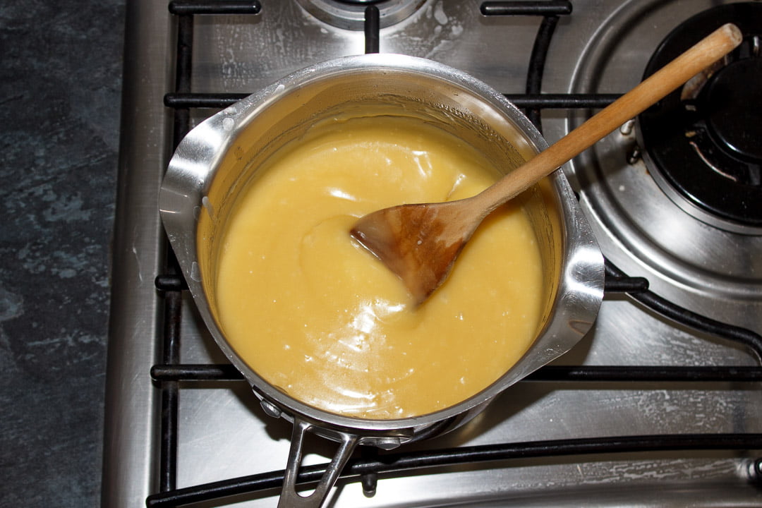 Melted butterscotch tart filling in a large saucepan on a stove top being beaten by a wooden spoon
