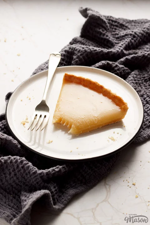 A slice of butterscotch tart on a plate with a fork, on a grey tea towel and white marble work top