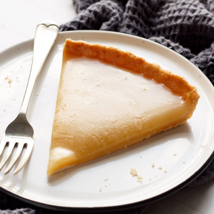 A slice of butterscotch tart on a plate with a fork, on a grey tea towel and white marble work top