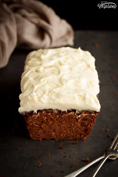 Carrot loaf cake topped with cream cheese frosting on a worktop with a cloth and forks in the background