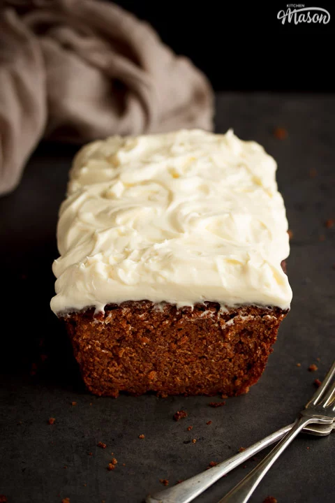 Carrot loaf cake topped with cream cheese frosting on a worktop with a cloth and forks in the background