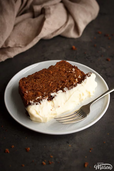 Slice of carrot loaf cake on a white plate with a fork and a cloth in the background