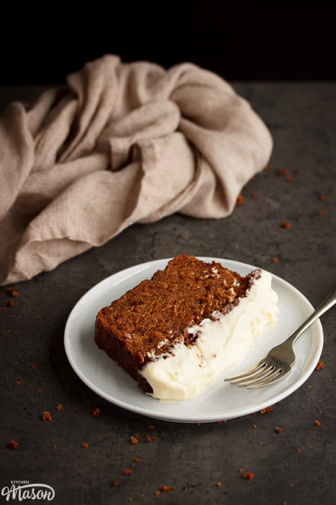 Slice of carrot loaf cake on a white plate with a fork and a cloth in the background