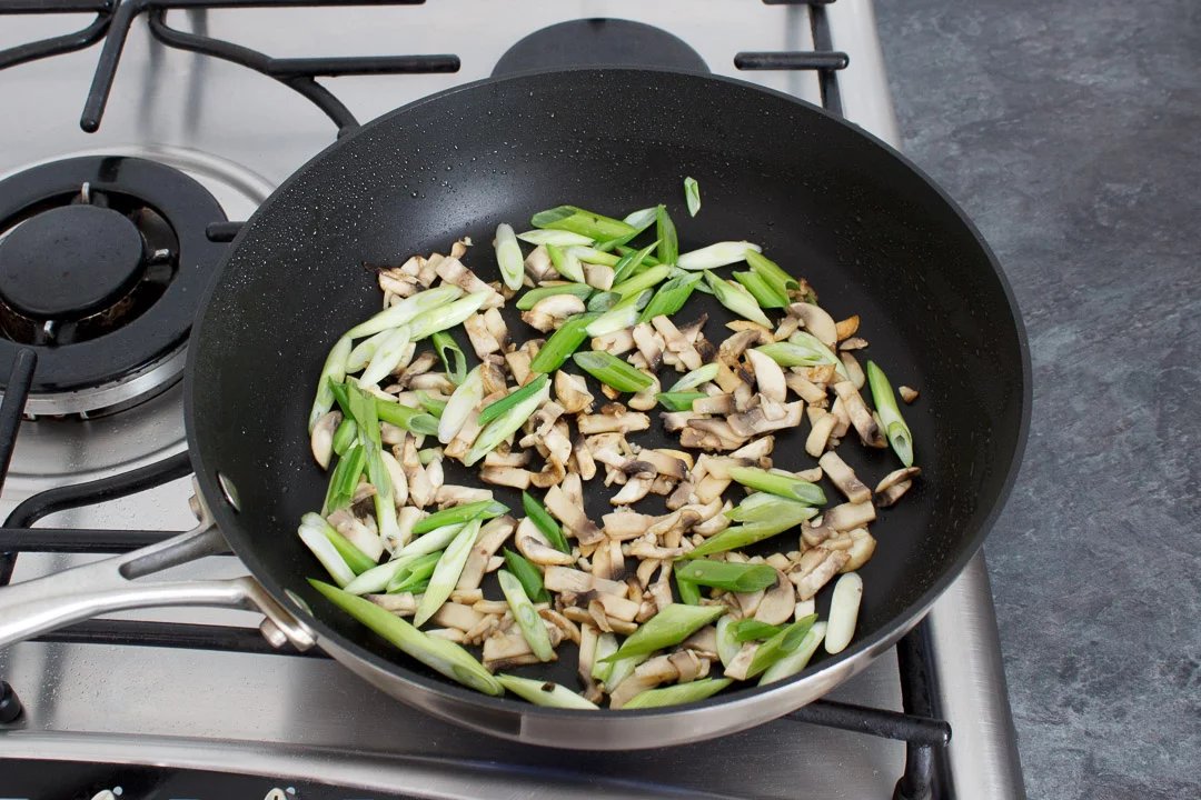 Chopped mushrooms and spring onion cooking a large frying pan on a hob