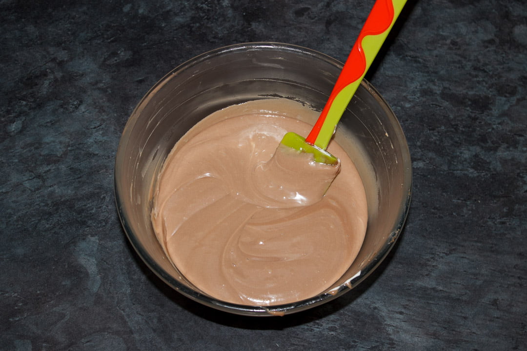 Whipped cream being folded into Kinder chocolate ganache in a glass bowl with a spatula