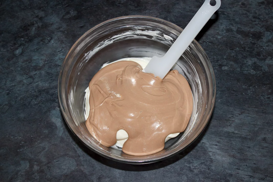 Kinder chocolate cream mixture being folded into the sweetened cream cheese mixture in a glass bowl with a spatula
