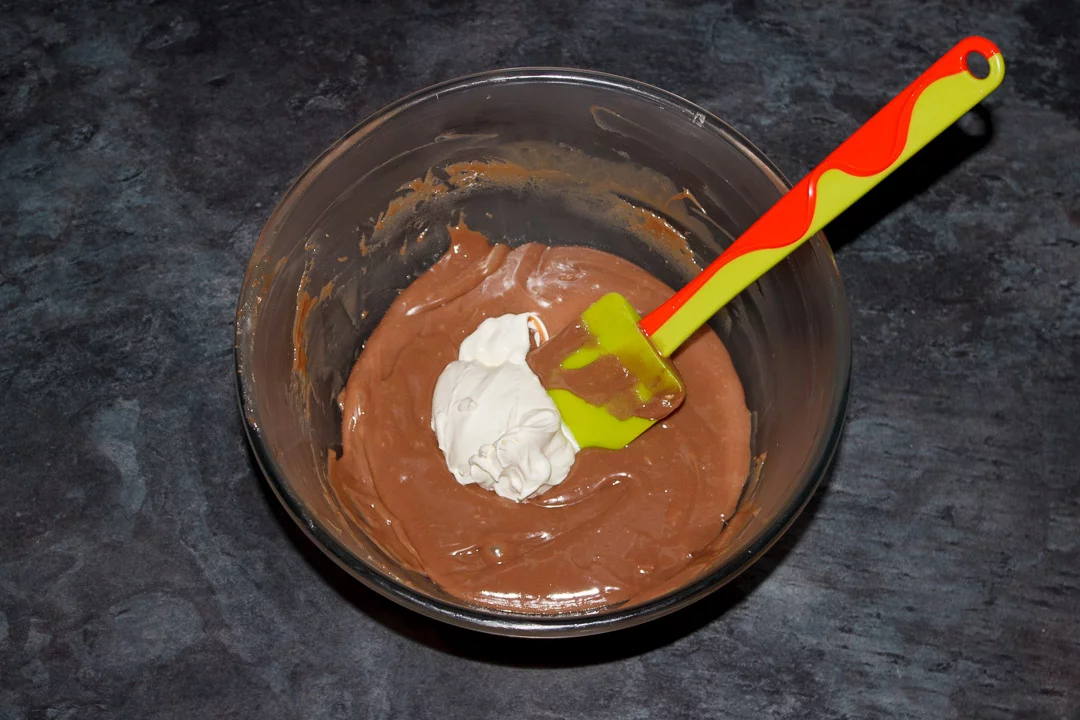 Whipped cream being folded into Kinder chocolate ganache in a glass bowl with a spatula