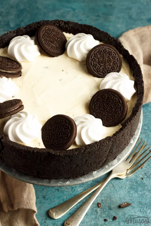 A whole no bake Oreo cheesecake on a cake stand with two forks, a light brown napkin and Oreo crumbs in the background