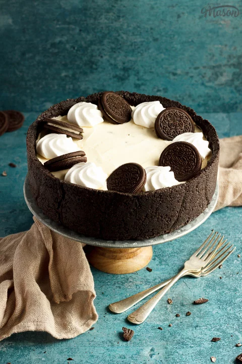 A whole no bake Oreo cheesecake on a cake stand with two forks, a light brown napkin and Oreo crumbs in the background