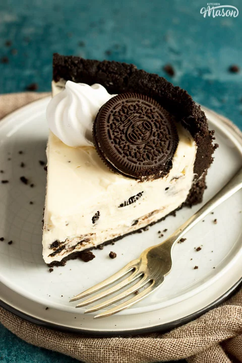 Slice of no bake Oreo cheesecake on a plate with a fork with a light brown napkin and Oreo crumbs in the background