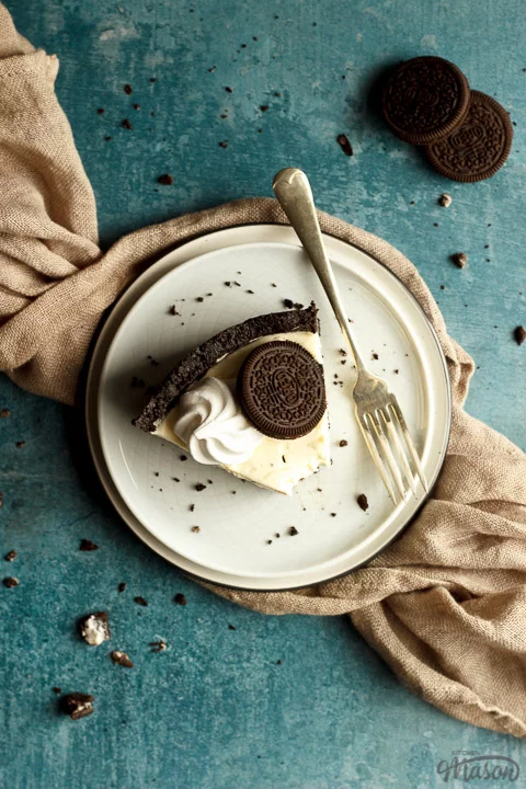 Slice of no bake Oreo cheesecake on a plate with a fork with a light brown napkin and Oreo crumbs in the background