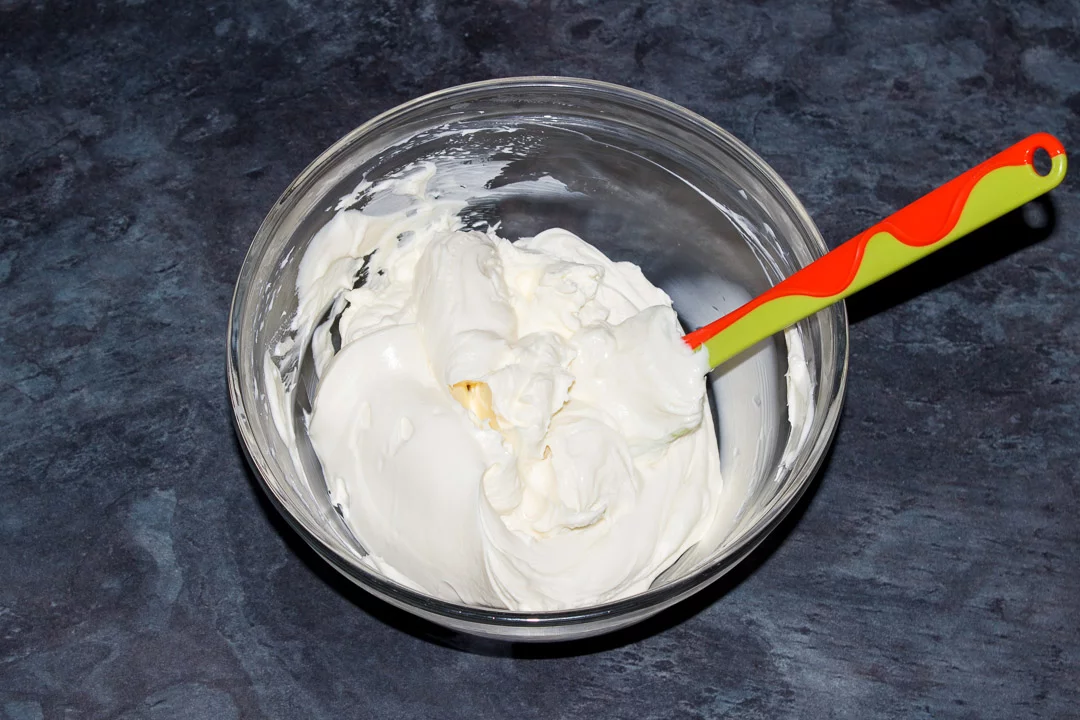 Whipped sweetened cream being folded into a cream cheese mixture with a rubber spatula in a glass bowl