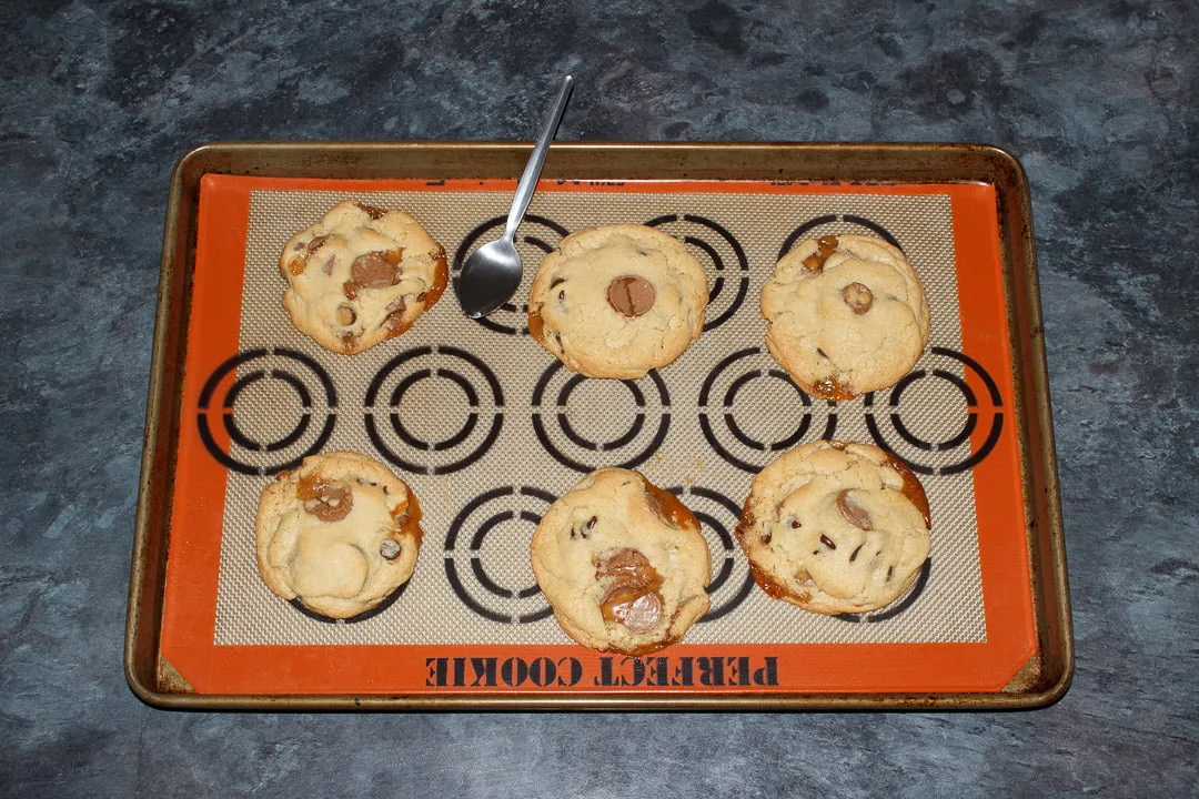 6 baked Rolo cookies spaced out on a large lined baking tray, oozing caramel has been pushed back into the cookies with a metal teaspoon