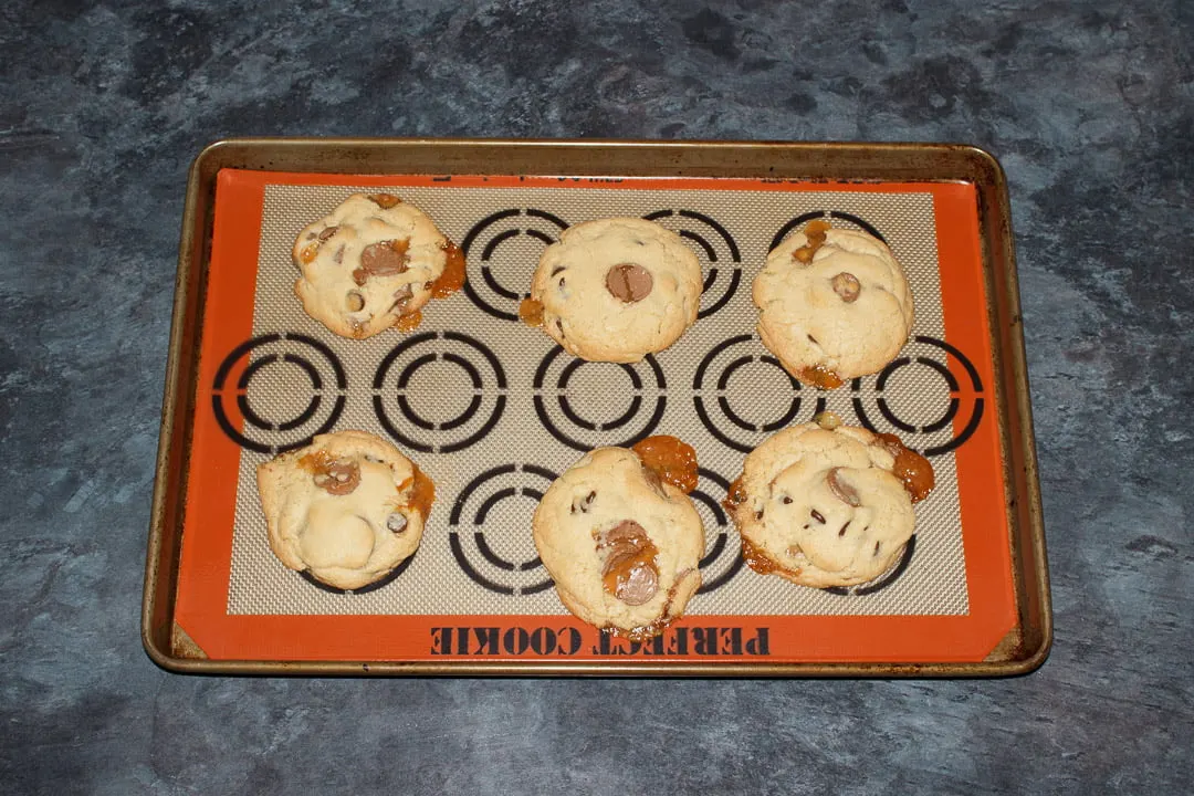 6 baked Rolo cookies spaced out on a large lined baking tray with caramel oozing out