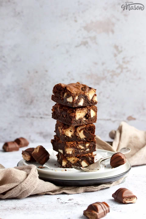 Kinder Bueno Brownies in a stack of 5 on a plate with a fork and 3 pieces of Kinder Bueno. With a light brown napkin and more Kinder Bueno pieces in the background.
