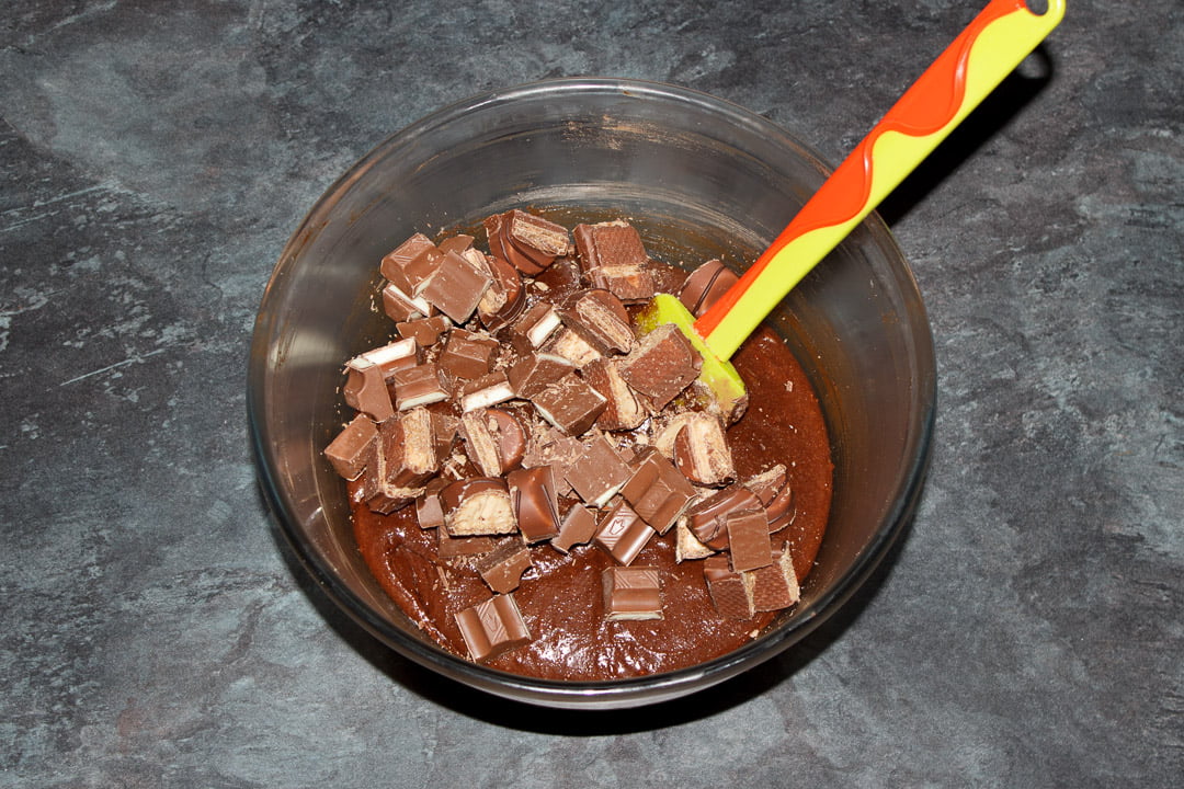 Brownie batter with pieces of chopped Kinder Bueno Bars and chopped Kinder Chocolate Bars on top in a glass bowl with a green spatula on top