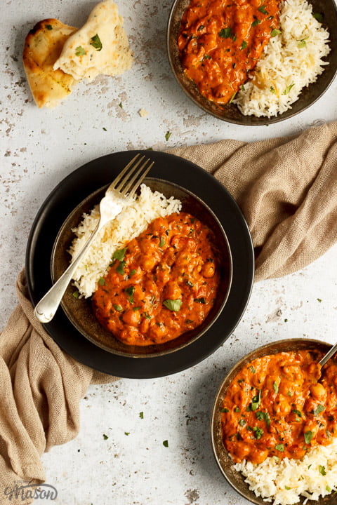 3 bowls of chana masala curry and rice on a textured white background with forks and a light brown napkin