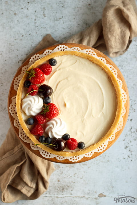 A no bake vanilla cheesecake on a cake stand topped with mixed berries surrounded by a light brown napkin