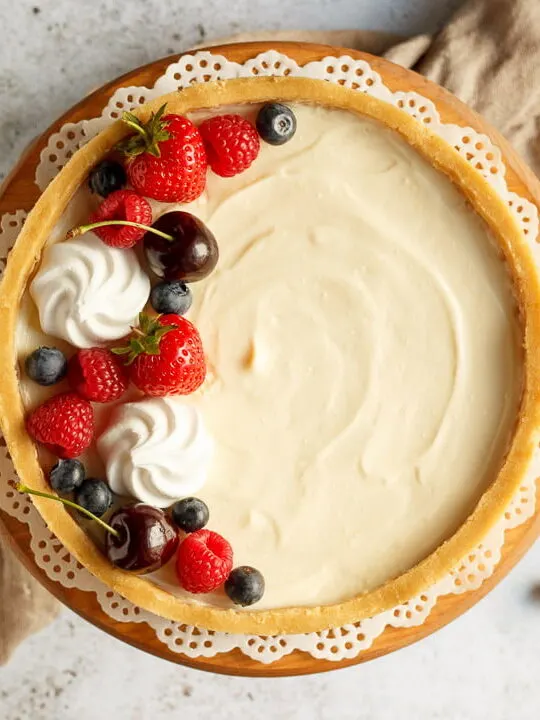 A no bake vanilla cheesecake on a cake stand topped with mixed berries surrounded by a light brown napkin