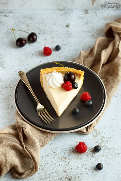 A slice of no bake vanilla cheesecake on a grey plate with a fork, topped with mixed berries, surrounded by a light brown napkin
