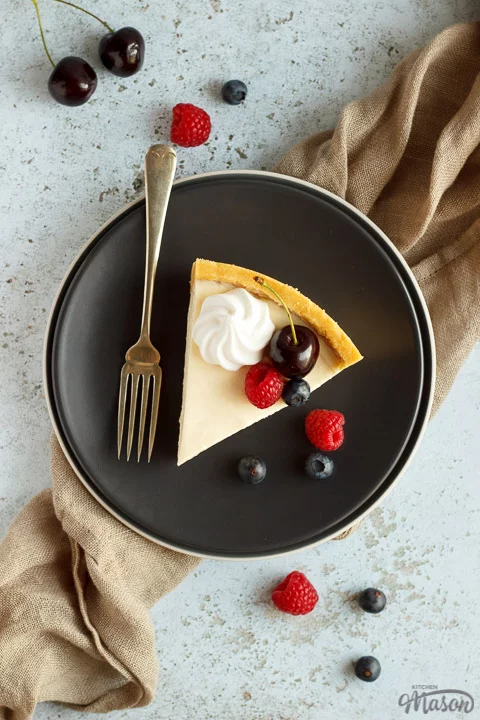 A slice of no bake vanilla cheesecake on a grey plate with a fork, topped with mixed berries, surrounded by a light brown napkin