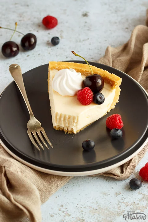 A slice of no bake vanilla cheesecake with a bite taken out of it on a grey plate with a fork, topped with mixed berries, surrounded by a light brown napkin