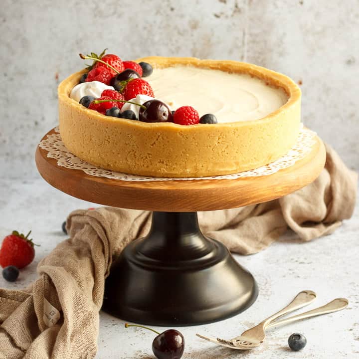 No bake vanilla cheesecake on a cake stand topped with mixed berries, surrounded by a light brown napkin and two forks
