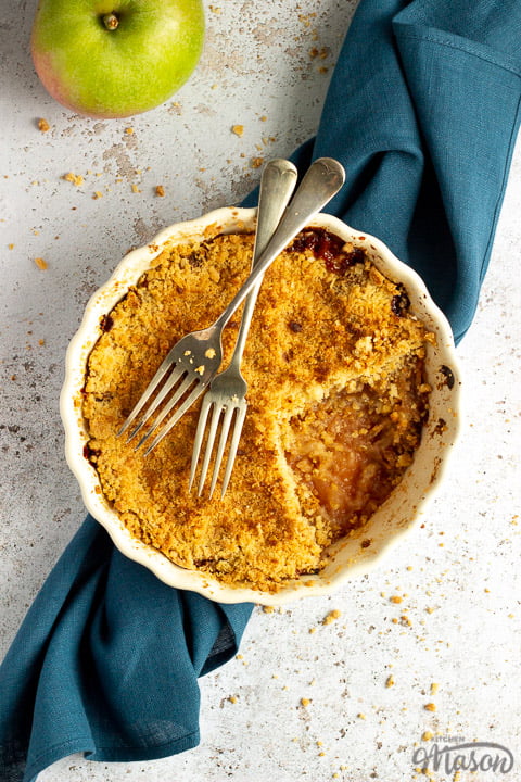 Apple crumble in a dish with two forks rested on top set over a blue linen napkin with a cooking apple in the background.