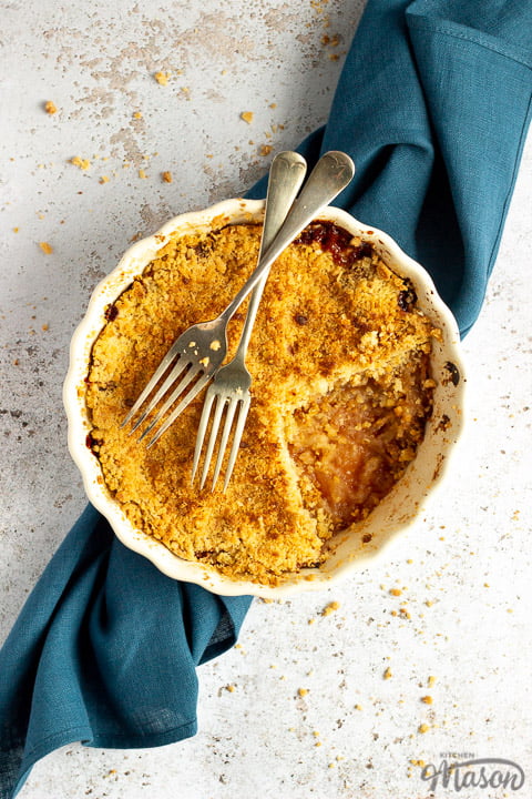 Apple crumble in a dish with two forks rested on top set over a blue linen napkin.