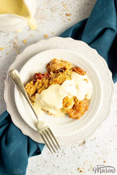 Slice of apple crumble in a small white bowl with a fork, drizzled with custard. There's a white jug of custard in the background and a blue linen napkin under the bowl.