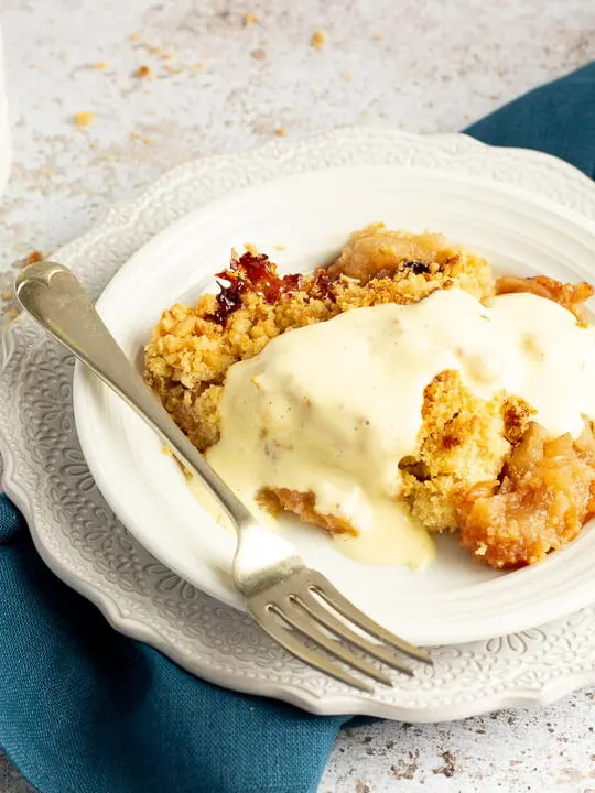 Slice of apple crumble in a small white bowl with a fork, drizzled with custard. There's a white jug of custard in the background and a blue linen napkin under the bowl.
