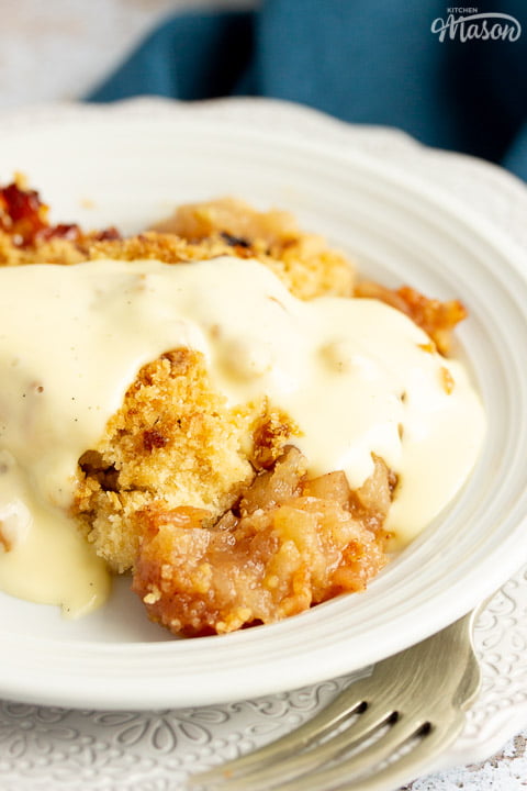 Slice of apple crumble in a small white bowl with a fork, drizzled with custard, set over a blue linen napkin.