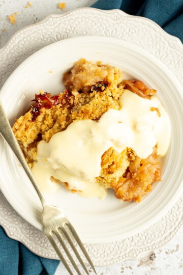 Slice of apple crumble in a small white bowl with a fork, drizzled with custard, set over a blue linen napkin.