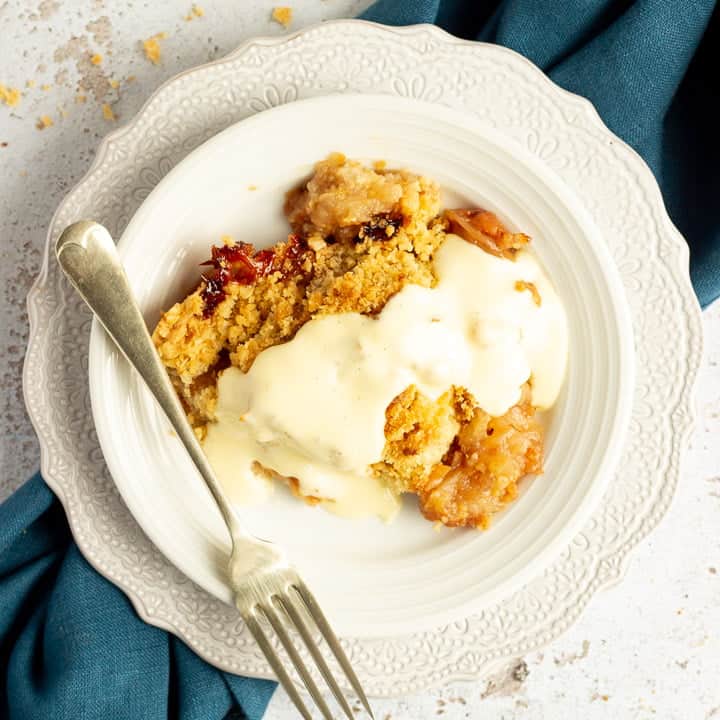 Slice of apple crumble in a small white bowl with a fork, drizzled with custard, set over a blue linen napkin.
