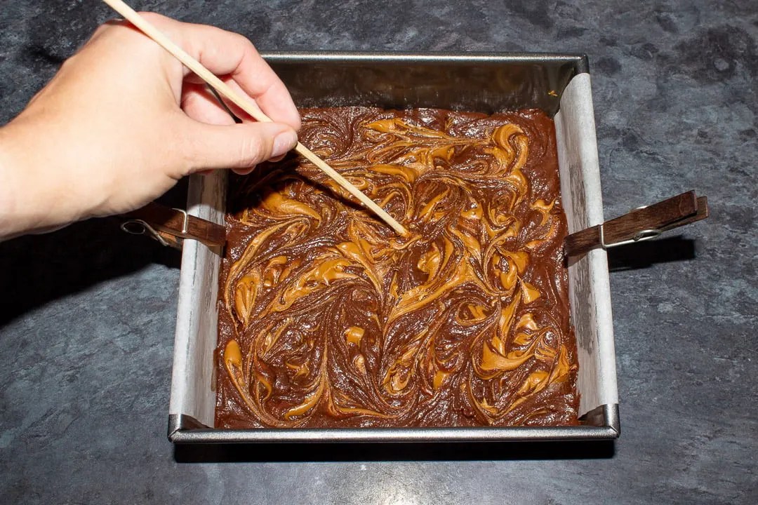 Biscoff spread being swirled into brownie batter with a skewer in a lined square baking tin