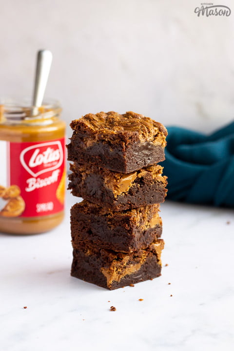 Biscoff brownies in a stack of 4 bars on a white work surface with a jar of Biscoff spread and a blue linen napkin in the background